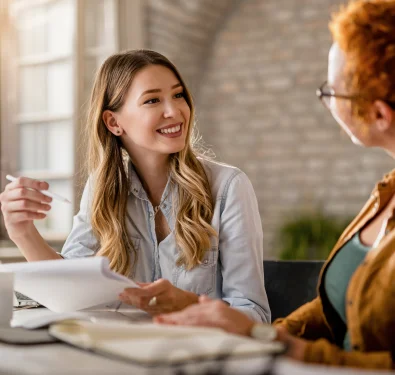 Een jonge blonde vrouw die enthousiast praat en documenten vasthoudt tijdens een gesprek met een collega in een creatieve studio.