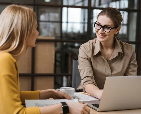 Een vriendelijke vrouw met een bril achter een laptop die glimlacht naar haar collega tijdens een zakelijke bijeenkomst.